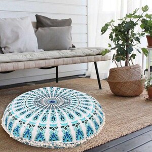 May include: Round floor cushion with a white and blue mandala design. The cushion has a white border with small, decorative tassels. The cushion is on a woven rug, with a bench and pillows in the background.