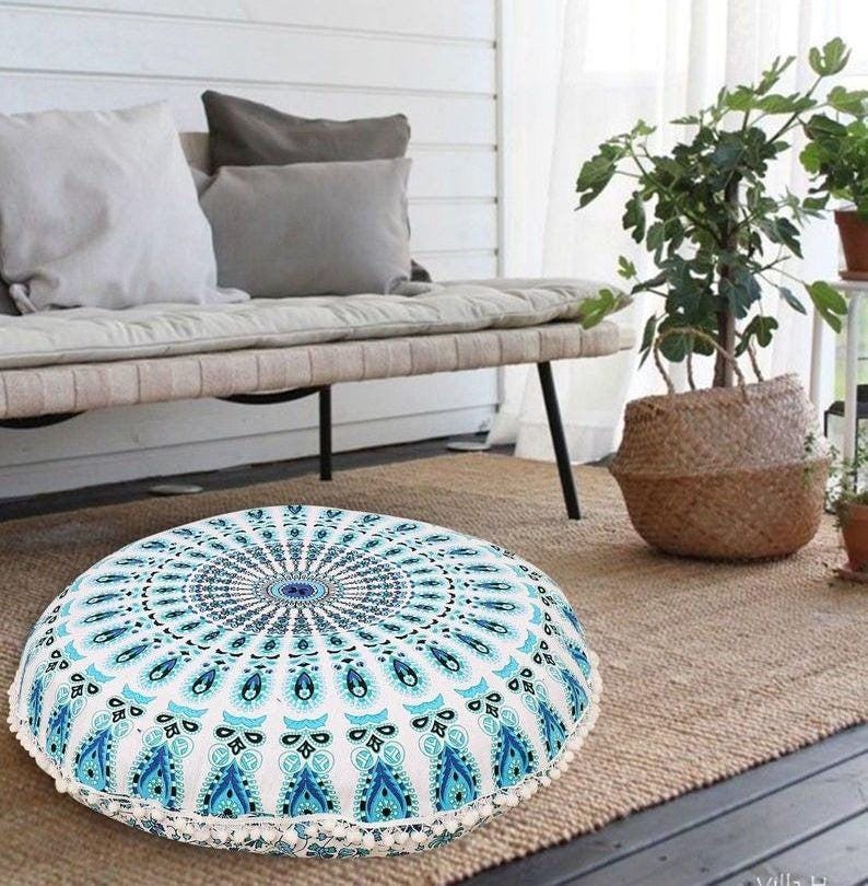 May include: Round floor cushion with a white and blue mandala design. The cushion has a white border with small, decorative tassels. The cushion is on a woven rug, with a bench and pillows in the background.