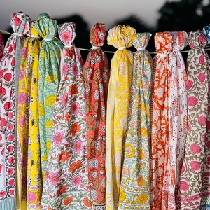 May include: A collection of colourful printed scarves with floral patterns hanging on a clothesline. The scarves are in shades of pink, yellow, green, orange, and blue.