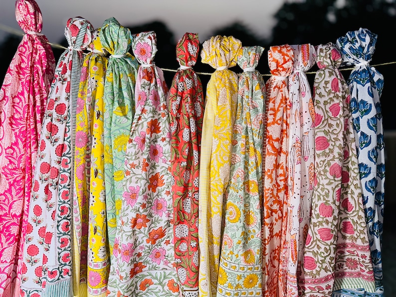May include: A collection of colourful printed scarves with floral patterns hanging on a clothesline. The scarves are in shades of pink, yellow, green, orange, and blue.