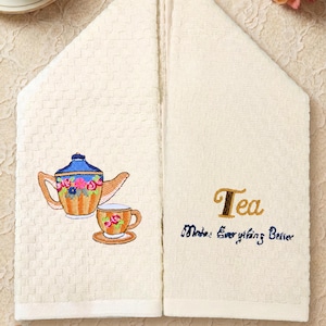 Puede incluir: Paño de cocina color crema con diseño bordado de tetera y taza. La toalla presenta la palabra "Tea" en escritura dorada, con la frase "Makes Everything Better" en escritura azul.