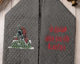 May include: A dark gray waffle-weave kitchen towel featuring an embroidered graphic of a horned figure in a red cloak standing on a green roof, with the red embroidered text "HE KNOWS WHEN YOU ARE SLEEPING" on the right side of the folded fabric.