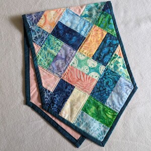 May include: A patchwork table runner with a diamond pattern in shades of blue, green, pink, and cream. The runner has a dark blue border.