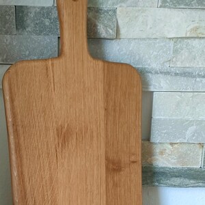 May include: A large, rectangular wooden cutting board with a handle. The board is made of light brown wood and has a natural finish. The board is sitting on a wooden surface.