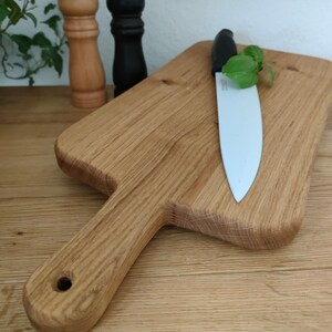 May include: A large wooden cutting board with a handle, a ceramic knife, and a sprig of green herbs. The board is made of light brown wood with a natural grain pattern.