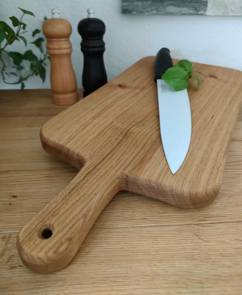May include: A large wooden cutting board with a handle, a ceramic knife, and a sprig of green herbs. The board is made of light brown wood with a natural grain pattern.