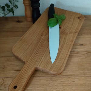 May include: A wooden cutting board with a handle, a white ceramic knife, and a sprig of green basil on top.
