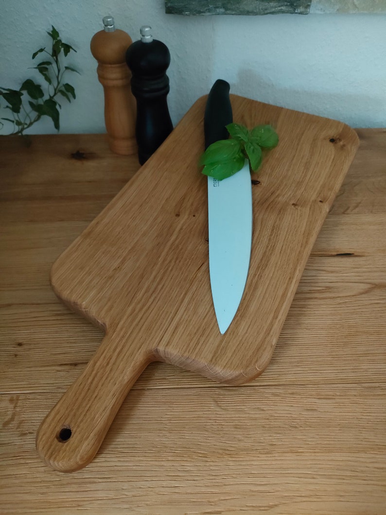 May include: A wooden cutting board with a handle, a white ceramic knife, and a sprig of green basil on top.