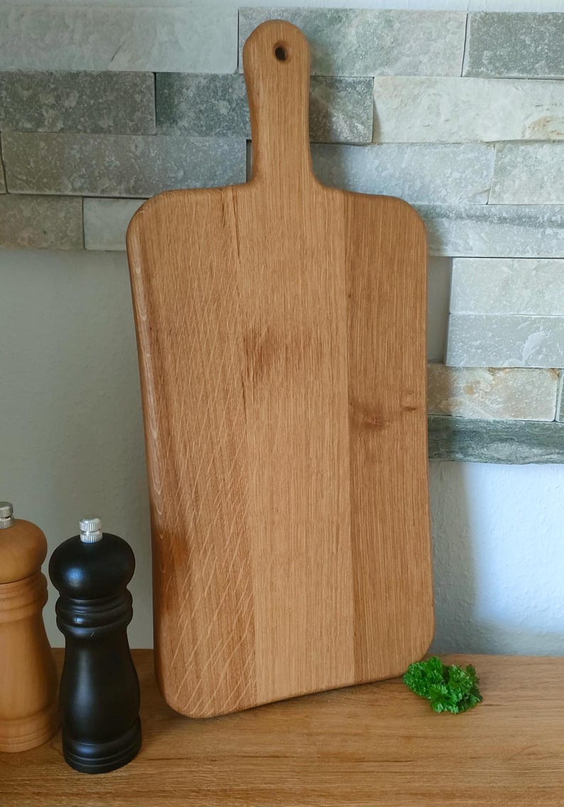 May include: A large, rectangular wooden cutting board with a handle. The board is made of light brown wood and has a natural finish. The board is sitting on a wooden surface.