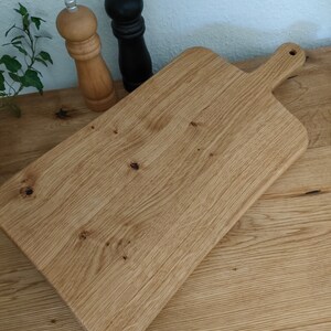 May include: A large, rectangular wooden cutting board with a handle. The board is made of light brown wood with a natural grain pattern. The board is sitting on a wooden surface.