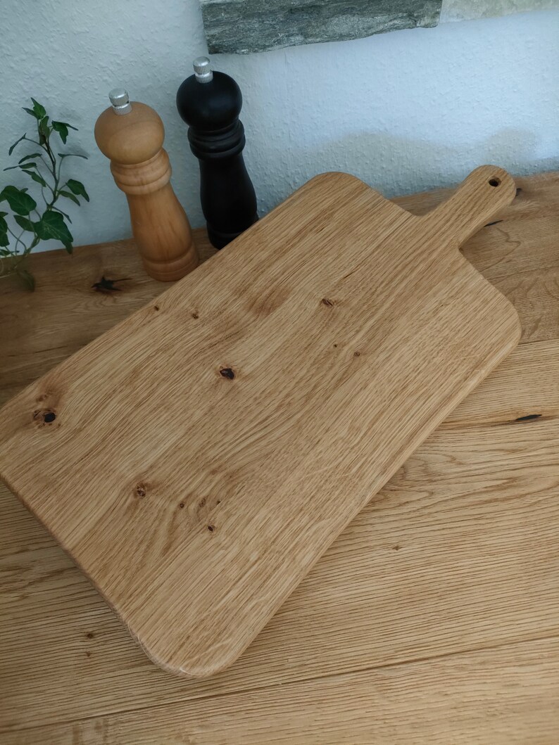 May include: A large, rectangular wooden cutting board with a handle. The board is made of light brown wood with a natural grain pattern. The board is sitting on a wooden surface.