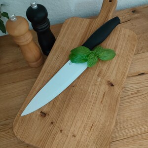 May include: A wooden cutting board with a white ceramic knife and a sprig of green basil on top. The board has a handle and is made of light brown wood.