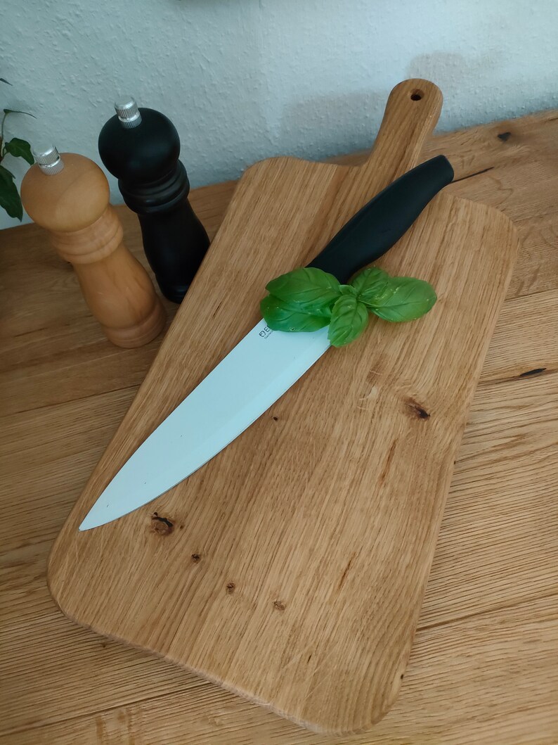 May include: A wooden cutting board with a white ceramic knife and a sprig of green basil on top. The board has a handle and is made of light brown wood.