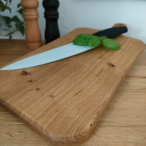 May include: A wooden cutting board with a light brown finish and a rounded edge. A white ceramic knife with a black handle is resting on the board, along with a sprig of green basil.