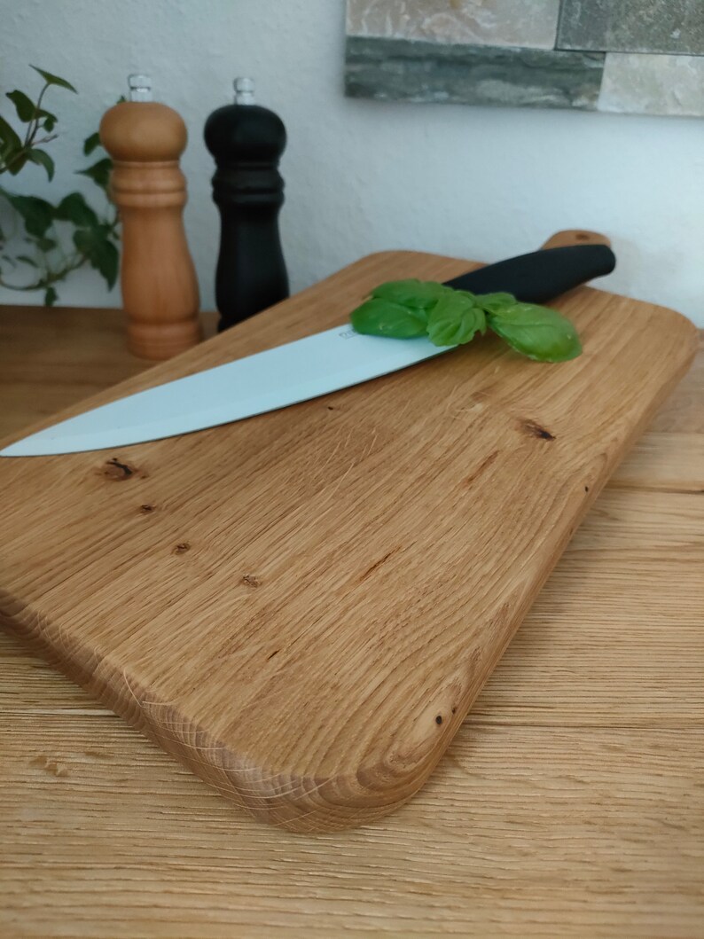 May include: A wooden cutting board with a light brown finish and a rounded edge. A white ceramic knife with a black handle is resting on the board, along with a sprig of green basil.