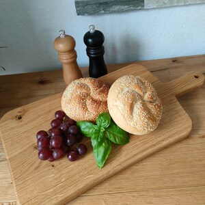 May include: A wooden cutting board with a handle, featuring two sesame seed rolls, a bunch of red grapes, and a sprig of green basil. Two salt and pepper shakers are visible in the background.
