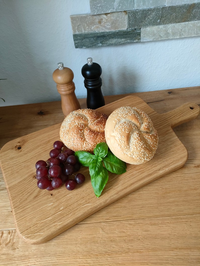 May include: A wooden cutting board with a handle, featuring two sesame seed rolls, a bunch of red grapes, and a sprig of green basil. Two salt and pepper shakers are visible in the background.