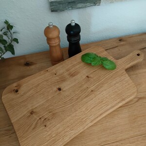 May include: A rectangular wooden cutting board with a handle on one side. The board is made of light brown wood and has a natural finish. There are two salt and pepper shakers on the counter behind the board, one black and one brown.