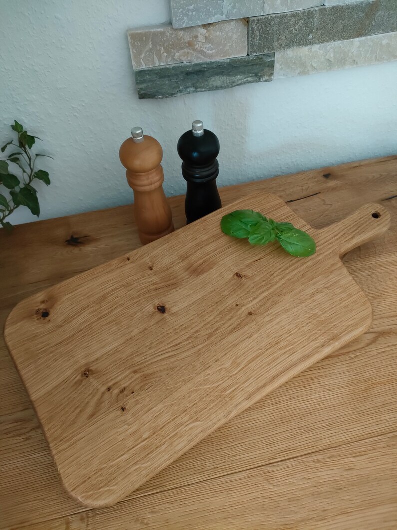 May include: A rectangular wooden cutting board with a handle on one side. The board is made of light brown wood and has a natural finish. There are two salt and pepper shakers on the counter behind the board, one black and one brown.