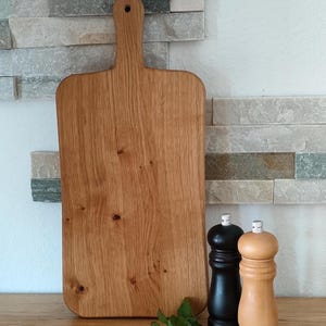 May include: A large, rectangular wooden cutting board with a handle. The board is made of light brown wood with a natural grain pattern. The board is sitting on a wooden surface next to two salt and pepper shakers.