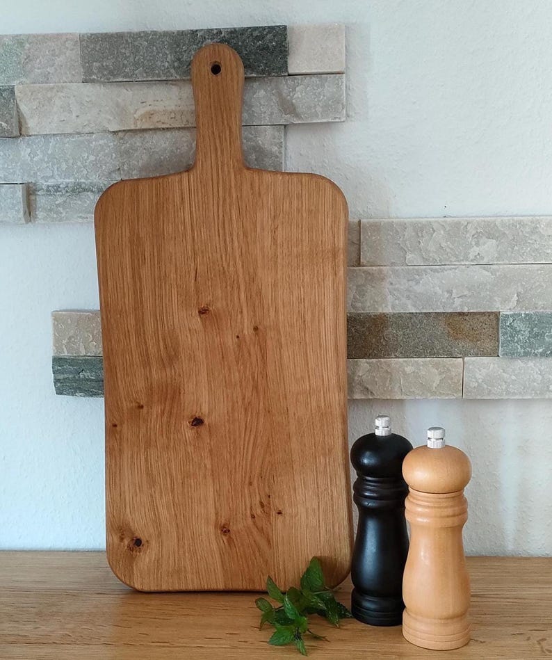 May include: A large, rectangular wooden cutting board with a handle. The board is made of light brown wood with a natural grain pattern. The board is sitting on a wooden surface next to two salt and pepper shakers.