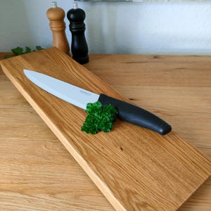 Può includere: Un tagliere in legno con un coltello a lama bianca e un rametto di prezzemolo. Il coltello ha un manico nero. Sullo sfondo ci sono due macinini di sale e pepe in legno. La tavola misura circa 40 cm di lunghezza.