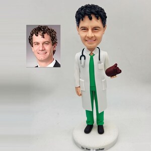 May include: A custom bobblehead figure of a doctor with dark curly hair, wearing a white lab coat over green scrubs, with a stethoscope around the neck and holding a dark red anatomical heart model. The base is inscribed with Dr. Rauscher.