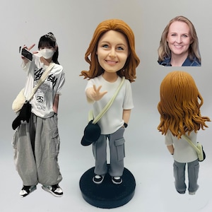 May include: A custom bobblehead figurine of a woman with red hair, wearing a white t-shirt, gray cargo pants, and a black baseball cap. The figurine is on a black base. A second person is also in the image, wearing a white t-shirt and gray pants.