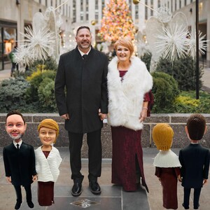 Op de afbeelding: Een stel staat hand in hand voor een kerstboom en feestelijke decoraties. De vrouw draagt een bordeauxrode jurk en een witte bontjas. De man draagt een zwarte jas. Miniatuurbeeldjes van een stel staan op de voorgrond.