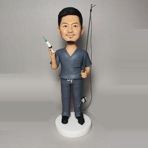 May include: A custom bobblehead figurine of a person wearing a blue medical scrub top and pants. The figurine is holding a syringe in one hand and a fishing rod in the other. The figurine is standing on a white base.