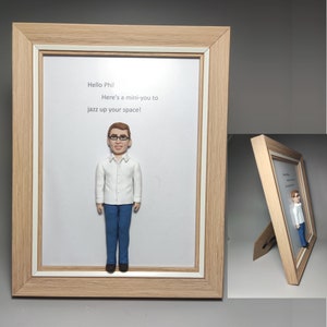 Può includere: Un ritratto incorniciato di una persona con occhiali, una camicia bianca e jeans blu. La cornice è in legno e ha un passepartout bianco. Il testo "Hello Phil Here's a mini-you to jazz up your space!" è stampato sul passepartout.
