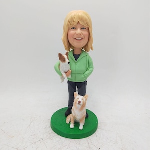 May include: A custom figurine of a woman with blonde hair wearing a green hooded sweatshirt. She is holding a small white and brown dog in her arms. A second dog, a white and brown dog with brown ears, is sitting on a green base.