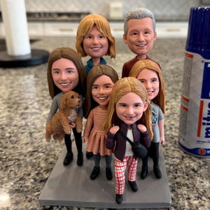Könnte beinhalten: Eine individuelle Bobblehead-Figur mit sieben Figuren, jeweils mit lächelndem Gesicht und detaillierter Kleidung. Eine Figur hält einen kleinen Hund. Die Figuren stehen auf einer grauen rechteckigen Basis, die ein Familien- oder Gruppenporträt darstellt.
