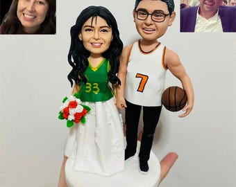 Op de afbeelding: Een op maat gemaakte bruidstaarttopper met een bruid in een groene en witte jurk met een boeket, en een bruidegom in een basketbalshirt met een bal. De topper bevat miniatuurportretten van het paar.