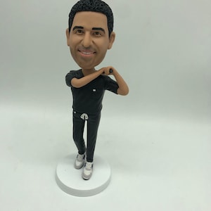 May include: A custom bobblehead figurine of a person wearing black scrubs and white trainers. The figurine is posed with one arm bent and the other arm extended.