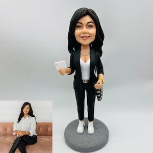 May include: A figurine of a woman in a black suit, white top, and white sneakers, holding a small white card. She has black hair and a purse with a black and white pattern. The figurine is on a gray circular base.