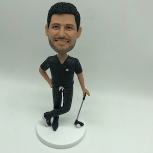 May include: A black and white custom bobblehead figurine of a person wearing a black scrub top and black trousers. The person is holding a golf club and a golf ball is on the ground.