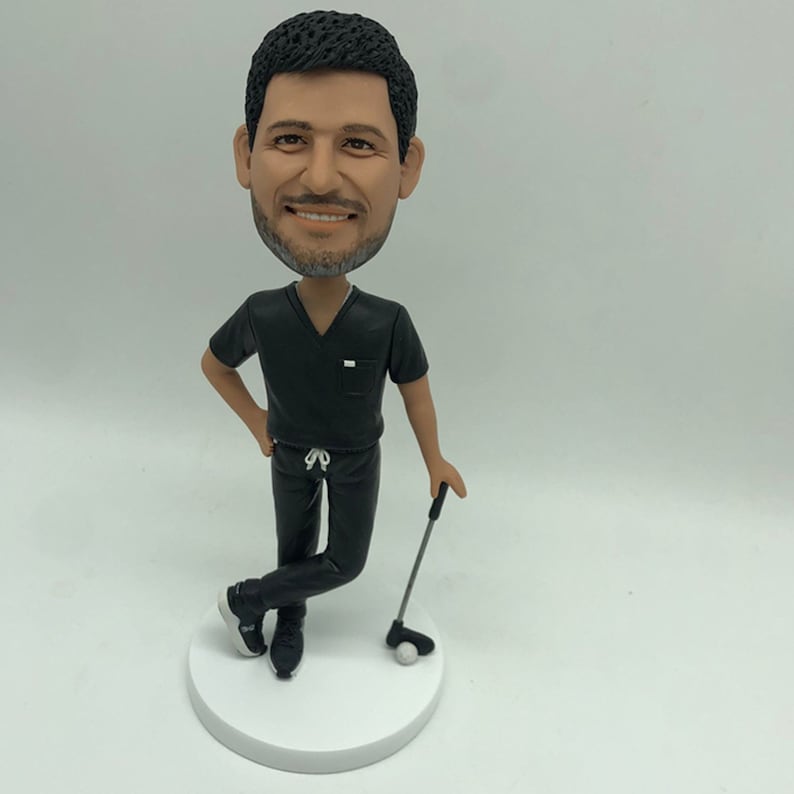 May include: A black and white custom bobblehead figurine of a person wearing a black scrub top and black trousers. The person is holding a golf club and a golf ball is on the ground.