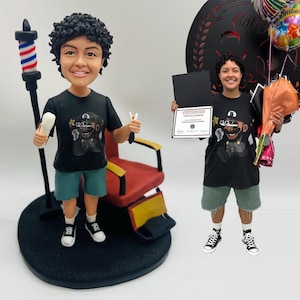 Op de afbeelding: Een beeldje van een persoon met krullend haar, gekleed in een zwart t-shirt met een grafisch ontwerp, groene shorts en zwart-witte sneakers. Het beeldje houdt kappersgereedschap vast en staat naast een kappersstoel en een barbierpaal. Het beeldje staat op een zwarte basis.