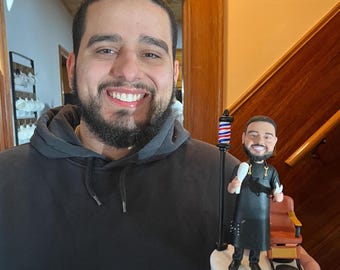 Puede incluir: Figura de cabeza oscilante de un barbero sosteniendo un peine y unas tijeras, de pie junto a un poste de barbero y una silla de barbero. La figura está sobre una base blanca. El barbero lleva un delantal negro.
