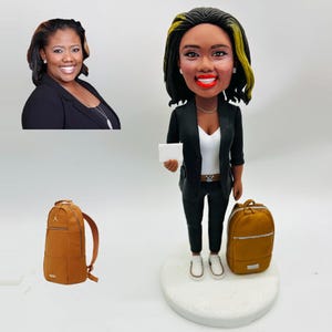 May include: A custom bobblehead of a woman in a black blazer, white top, and black pants, holding a small white card. A brown backpack and a photo of the woman are also in the image. The bobblehead is on a white base.