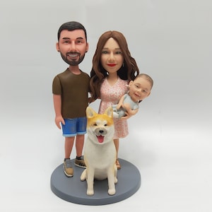 May include: A custom figurine set of a family with a dog. The figurines are made of painted ceramic and depict a man, woman, and child. The dog is a light brown and white Akita. The figurines are standing on a gray base.
