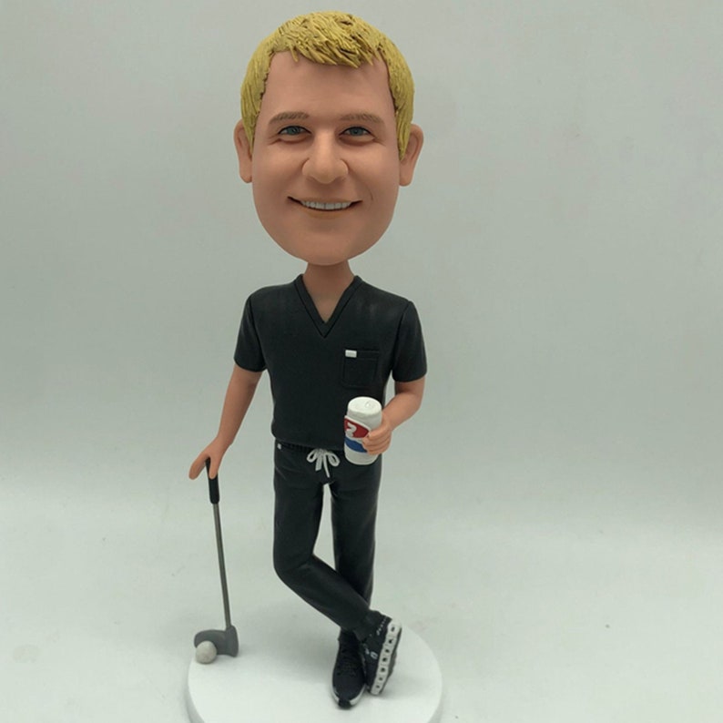 May include: A custom bobblehead figurine of a person wearing a black scrub top and black trousers. The person is holding a can of soda in one hand and a golf club in the other. The person has short blond hair and is smiling.