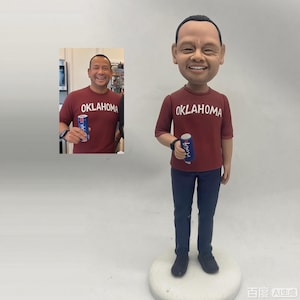 May include: A custom bobblehead figurine depicting a person with short dark hair, wearing a maroon t-shirt with the word OKLAHOMA printed in white across the chest, dark blue pants, and black shoes, holding a small blue beverage can in one hand.