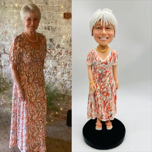 May include: A woman in a long, patterned dress stands next to a bobblehead figurine wearing a similar dress. The dress features a white background with red and brown abstract designs. The figurine has a smiling face and is on a black base.