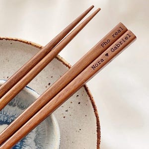Op de afbeelding: Houten eetstokjes met de tekst "Pho real" en "Nora ❤ Gabriel" gegraveerd. De eetstokjes liggen op een gespikkelde keramische plaat en kom. De eetstokjes zijn middelbruin.