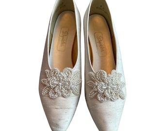 May include: A pair of off-white, pointed-toe pumps. Each shoe features a decorative floral embellishment with pearl-like beads. The shoes have a low heel and the brand name "Depides" is visible inside.