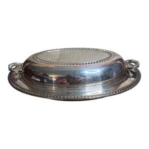 May include: A silver-colored, oval-shaped serving dish with a lid. The dish has a decorative beaded edge and loop handles on either side. The lid is also oval and has a similar beaded edge. The dish sits on a round, silver-colored base.