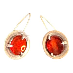 May include: A pair of silver-toned earrings. Each earring features an oval, reddish-orange stone set within a silver frame. The earrings have a simple, elegant design with a hook closure.