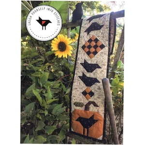 May include: A black and orange Halloween table runner with a pumpkin, crows, and a checkered pattern. The table runner is hanging on a wooden fence with a sunflower in the background. The text "Laugh Yourself Into Stitches" is visible in a circle.